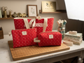 Heritage Homestead Quilted Travel Bundle featuring a matching red and cream calico canvas tote bag with a cream bow and lace trim, a boxy cosmetic case, and a flat zipper pouch on a wooden cutting board.