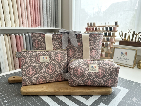 Vintage Damask Quilted Travel Bundle featuring a matching charcoal grey and blush pink floral baroque canvas tote bag with a polka dot bow, a boxy cosmetic case, and a flat zipper pouch on a wooden board.