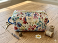 Cottage Garden Quilted Makeup Bag canvas cosmetic case featuring a white floral watercolor print, a blue fabric grab handle, and a top zipper on a wooden table next to thread spools and a thimble.
