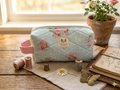 Pastel Rose and Polka Dot Quilted Cosmetic Case canvas makeup bag featuring a light blue, white dot, and pink rose print, a pink fabric grab handle, and a top zipper on a rustic wooden table next to vintage thread spools and a potted rose.