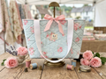 Pastel Rose & Polka Dot quilted canvas tote bag featuring a light blue background with white polka dots and pink roses, a front pocket with brand label, wide cream shoulder straps, and a pink ribbon bow closure.