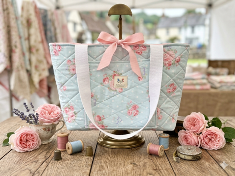 Pastel Rose & Polka Dot quilted canvas tote bag featuring a light blue background with white polka dots and pink roses, a front pocket with brand label, wide cream shoulder straps, and a pink ribbon bow closure.
