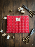 Heritage Homestead Quilted Wallet canvas zipper pouch featuring a red and cream calico floral print, a cream fabric attachment loop, and a gold swivel clip flat on a dark wooden table surrounded by vintage scissors and thread spools.