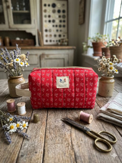 Heritage Homestead Quilted Makeup Bag canvas cosmetic case featuring a red and cream calico floral print, a cream fabric side handle, and gold top zipper on a rustic wooden table next to antique scissors and thread spools.