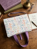 Joyful Grace Script Quilted Premium Bookcover featured in FB_IMG_1783682781995.jpg showcasing a padded Bible sleeve with colorful faith, hope, and love script, purple floral handles, shoulder strap, and ribbon marker on a wooden table.