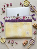 A yellow and white gingham laptop sleeve with a purple ruffled trim and a floral patterned band, shown holding a silver laptop. The sleeve is styled in a flat-lay arrangement with paintbrushes, paint palettes, and small pink flowers on a beige fabric background.