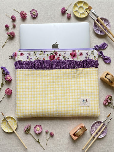 A yellow and white gingham laptop sleeve with a purple ruffled trim and a floral patterned band, shown holding a silver laptop. The sleeve is styled in a flat-lay arrangement with paintbrushes, paint palettes, and small pink flowers on a beige fabric background.