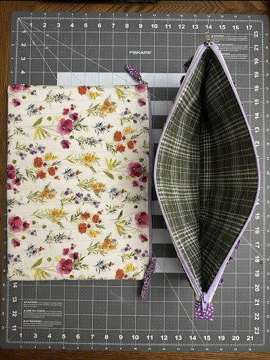 A fabric zippered pouch shown open next to its floral-patterned cover on a gray cutting mat. The cover has a cream background with a multicolor floral print, and the open pouch shows a green plaid lining with lavender trim and matching lavender wristlets.