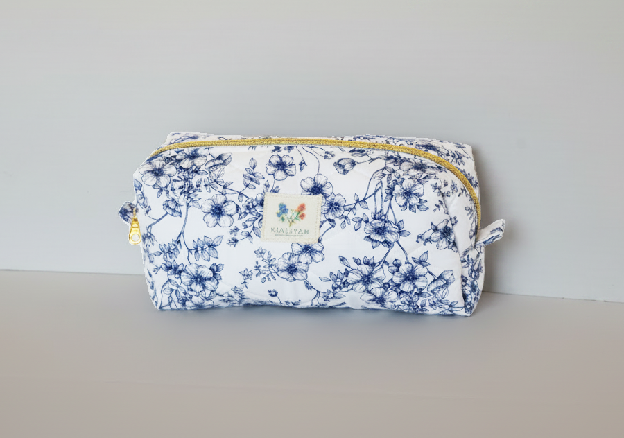 The Blue Floral Quilted Toile Makeup Bag featuring a monochrome blue pastoral toile print on cotton fabric, designed with vertical channel quilting, a flat stand-up base, and a gold-tone zipper with a matching fabric pull-tab.