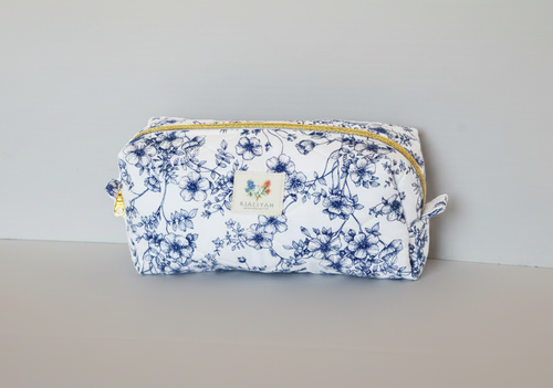 The Blue Floral Quilted Toile Makeup Bag featuring a monochrome blue pastoral toile print on cotton fabric, designed with vertical channel quilting, a flat stand-up base, and a gold-tone zipper with a matching fabric pull-tab.