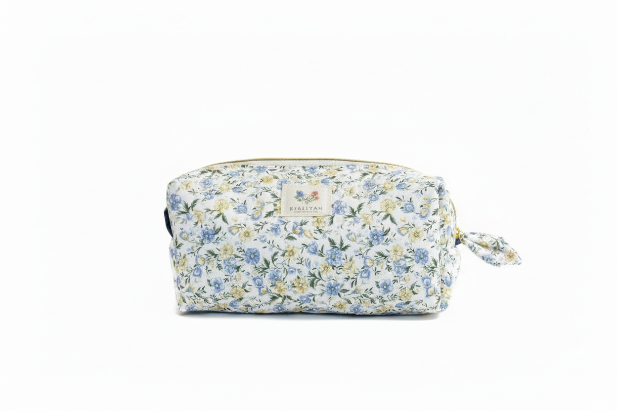 The Meadow Flora Quilted Cosmetic Pouch featuring a delicate blue and yellow botanical print on cream fabric, designed with vertical channel quilting, a flat stand-up base, a gold-tone zipper with a matching fabric pull-tab, and a centered white KIALIYAH label.