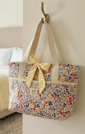 Prairie Wildflower Quilted Tote showcasing an oversized colorful floral calico handbag with horizontal quilting, natural canvas shoulder straps, and a yellow gingham check bow hanging on a brass wall hook.