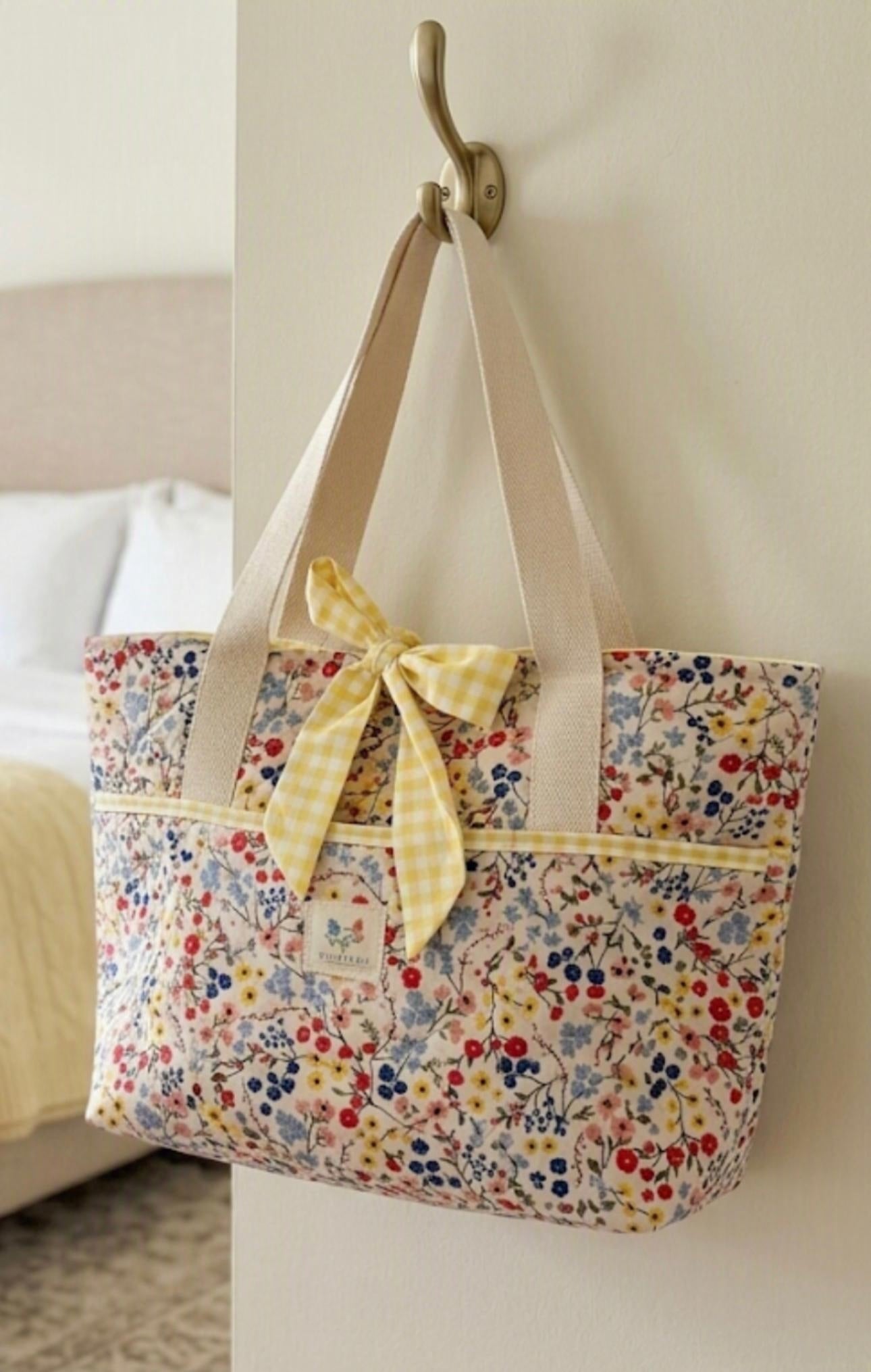 Prairie Wildflower Quilted Tote showcasing an oversized colorful floral calico handbag with horizontal quilting, natural canvas shoulder straps, and a yellow gingham check bow hanging on a brass wall hook.