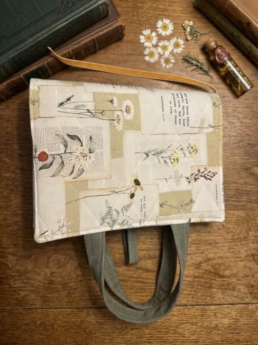 Pressed Floral Parchment Quilted Premium Bookcover showcasing a padded Bible sleeve with pressed daisy and vintage book page print, sage green handles, and a gold ribbon bookmark on a wood table.