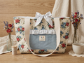 Handcrafted 3-pocket diamond-quilted tote bag by Kialiyah featuring a red rose and blue floral pattern, a light blue chambray front pocket with ruffled trim, a matching top fabric tie bow, and cream webbed shoulder straps.