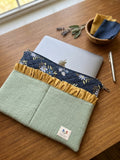 A sage green quilted laptop sleeve with a mustard yellow ruffle and a navy floral patterned top section, shown on a wooden desk with a silver laptop, a pen, and a small bowl of cloths.