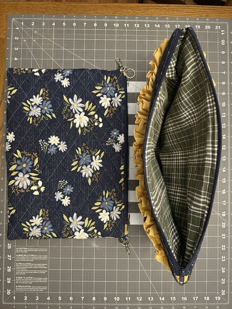 A handmade fabric pouch shown on a gray cutting mat, displaying both the floral exterior (navy blue with white and yellow flowers) and the interior (green plaid lining with a mustard yellow ruffled trim).