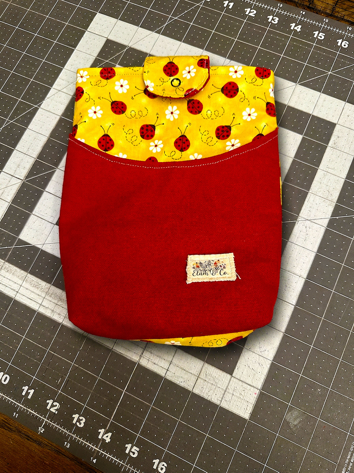 Handcrafted tablet sleeve featuring a solid red canvas front pocket, a sunny yellow upper panel printed with ladybugs and daisies, a matching snap closure tab, and an Elam & Co. fabric label resting on a cutting mat.