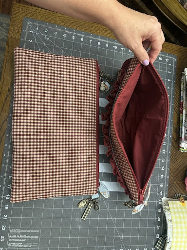A handmade fabric pouch with a brown and beige gingham patterned exterior and a deep burgundy interior, shown on a gray cutting mat with a hand holding the open zipper.