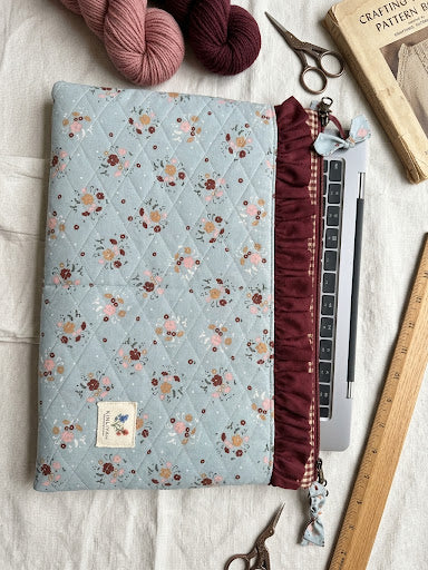 A quilted fabric laptop sleeve with a light blue floral pattern and a burgundy ruffled zipper, shown on a white fabric background with yarn, scissors, a book, and a wooden ruler.