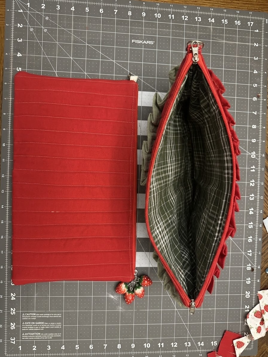A red fabric zippered pouch shown closed and open on a gray measuring mat. The closed pouch is plain red with subtle vertical stitched lines, and the open pouch reveals a gray plaid lining and red ruffled trim along the edges.