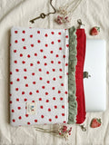 A quilted fabric laptop sleeve with a white background, red strawberry pattern, and a red ruffled zipper, shown on a beige linen background with a silver laptop, decorative key, and dried flowers.