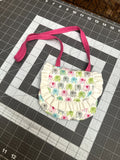 Handcrafted toddler purse made from white fabric featuring a colorful pastel elephant print, a cream ruffled edge flap, and a bright pink shoulder strap resting on a cutting mat.