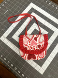 Handcrafted fabric toddler purse printed with red, pink, and white patterned hearts, featuring a red ruffled edge flap and matching red crossbody strap on a cutting grid.