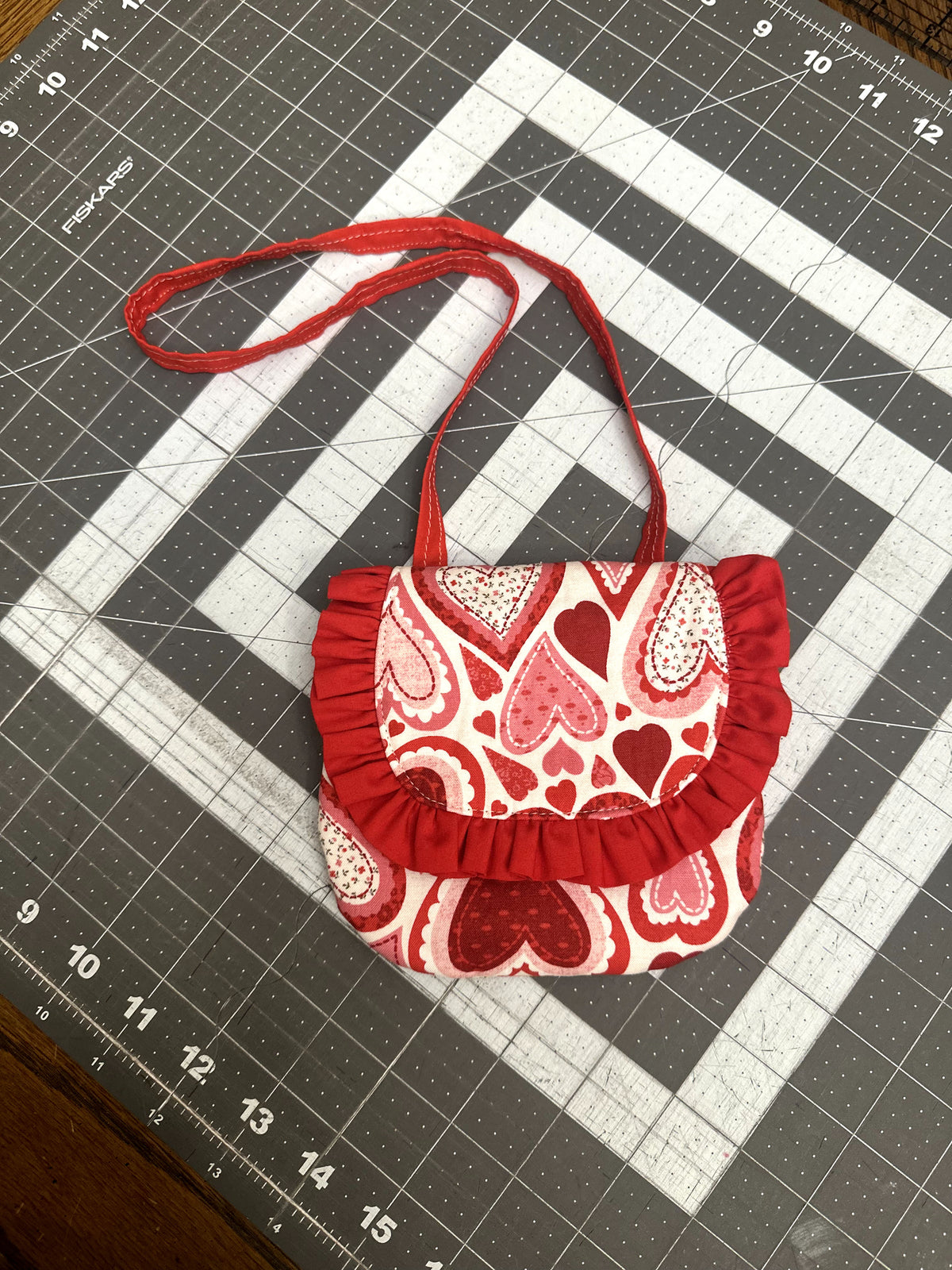 Handcrafted fabric toddler purse printed with red, pink, and white patterned hearts, featuring a red ruffled edge flap and matching red crossbody strap on a cutting grid.