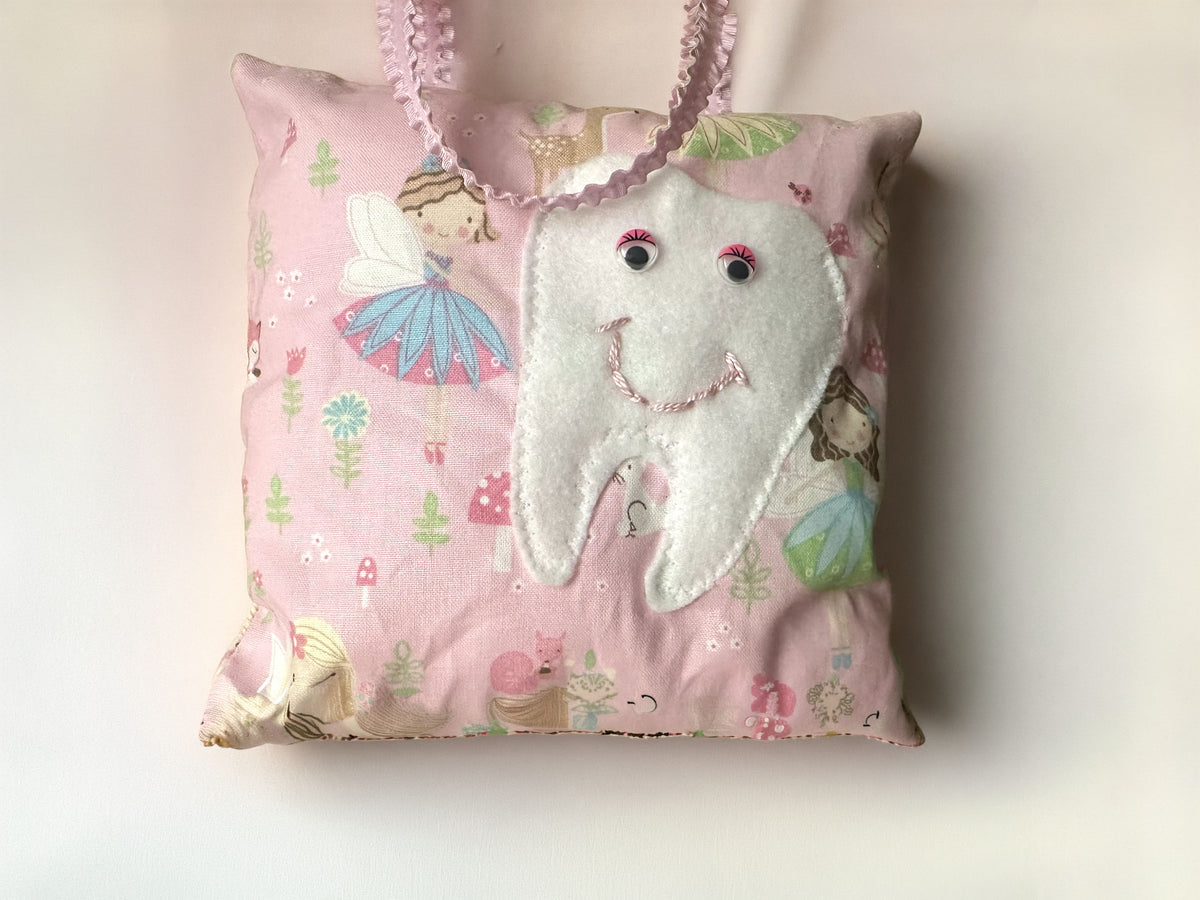 Handcrafted pink fairy-themed tooth pillow featuring magical garden fairies, a soft felt tooth pocket with pink eyelashes, and a ruffled pink hanging ribbon handle on a white background.