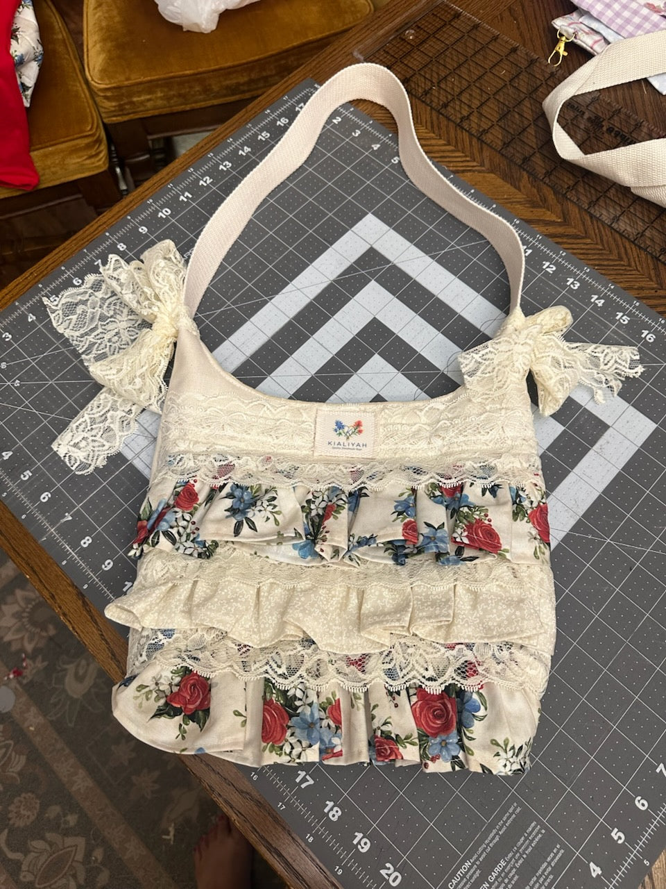 Vintage Rose Tiered Ruffle Purse showcasing a handmade cream shoulder bag with alternating tiers of red and blue rose print ruffles, detailed with wide eyelet lace borders and two large lace bows on a rustic wooden table.