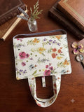 Wildflower Meadow Quilted Premium Bookcover showcasing a cream padded book sleeve with watercolor poppy and lavender botanical print, matching handles, and a lavender velvet ribbon bookmark on a wood table.