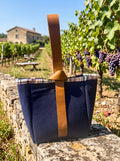 Large Navy Flannel-Rimmed Wine Tote featured in Large Wine Bag.png showcasing a deep navy canvas bottle carrier with a plaid flannel rim and a rich caramel leather loop-knot handle sitting on a stone wall with a vineyard background.