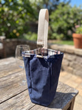 Medium Navy Flannel-Rimmed Wine Tote featured in Medium Wine Bag.png showcasing a single-bottle navy canvas carrier with a plaid flannel rim and a natural cotton webbing strap clipped to silver metal rings on a rustic wooden table.
