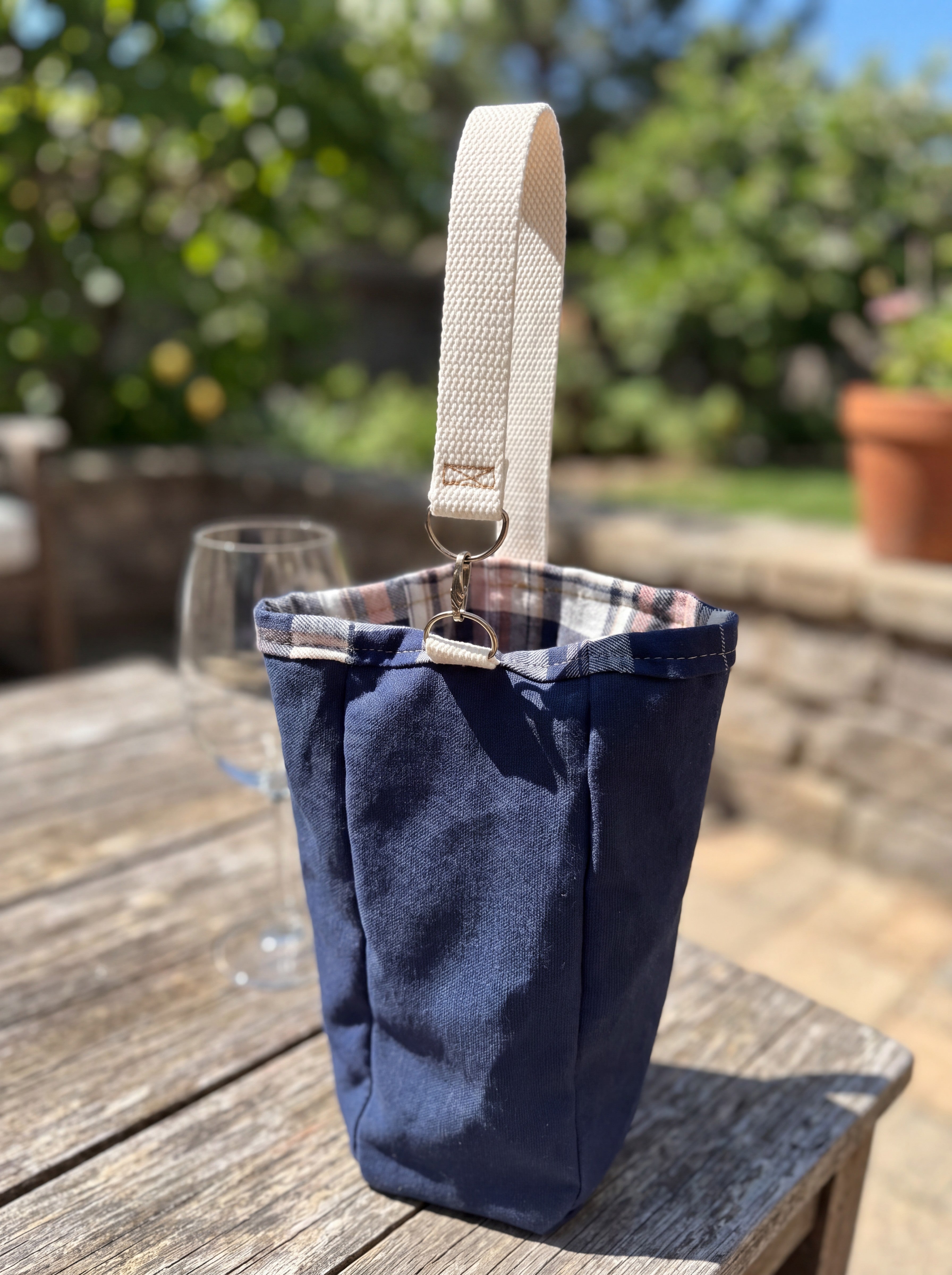 Medium Navy Flannel-Rimmed Wine Tote featured in Medium Wine Bag.png showcasing a single-bottle navy canvas carrier with a plaid flannel rim and a natural cotton webbing strap clipped to silver metal rings on a rustic wooden table.