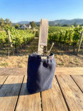 Small Navy Flannel-Rimmed Wine Tote featured in Small Wine Bag.png showcasing a compact single-bottle navy canvas pouch with a plaid flannel rim and a cream cotton webbing handle clipped to metal hardware rings resting on a wooden slatted table with a sunny vineyard landscape in the background.