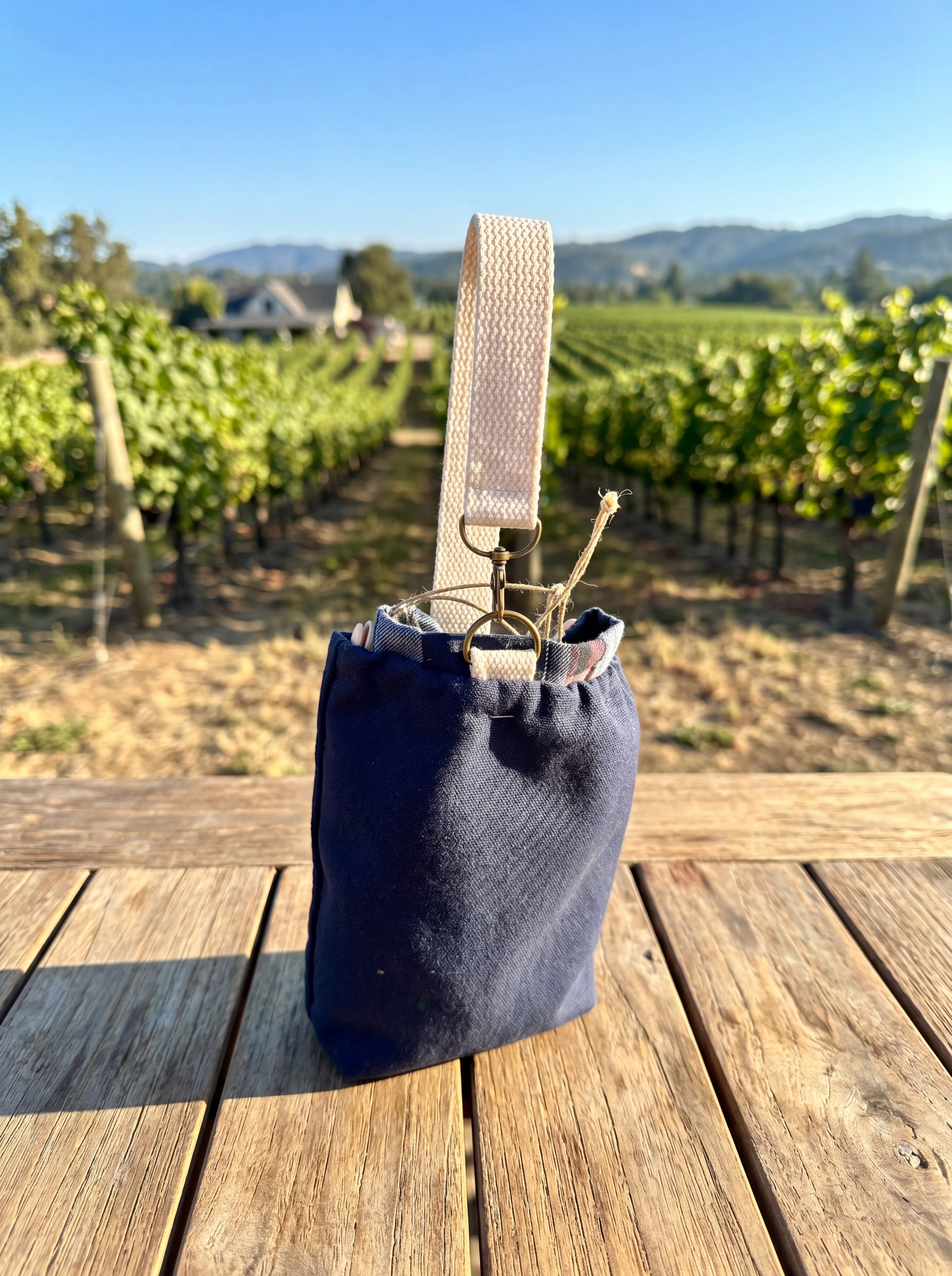 Small Navy Flannel-Rimmed Wine Tote featured in Small Wine Bag.png showcasing a compact single-bottle navy canvas pouch with a plaid flannel rim and a cream cotton webbing handle clipped to metal hardware rings resting on a wooden slatted table with a sunny vineyard landscape in the background.