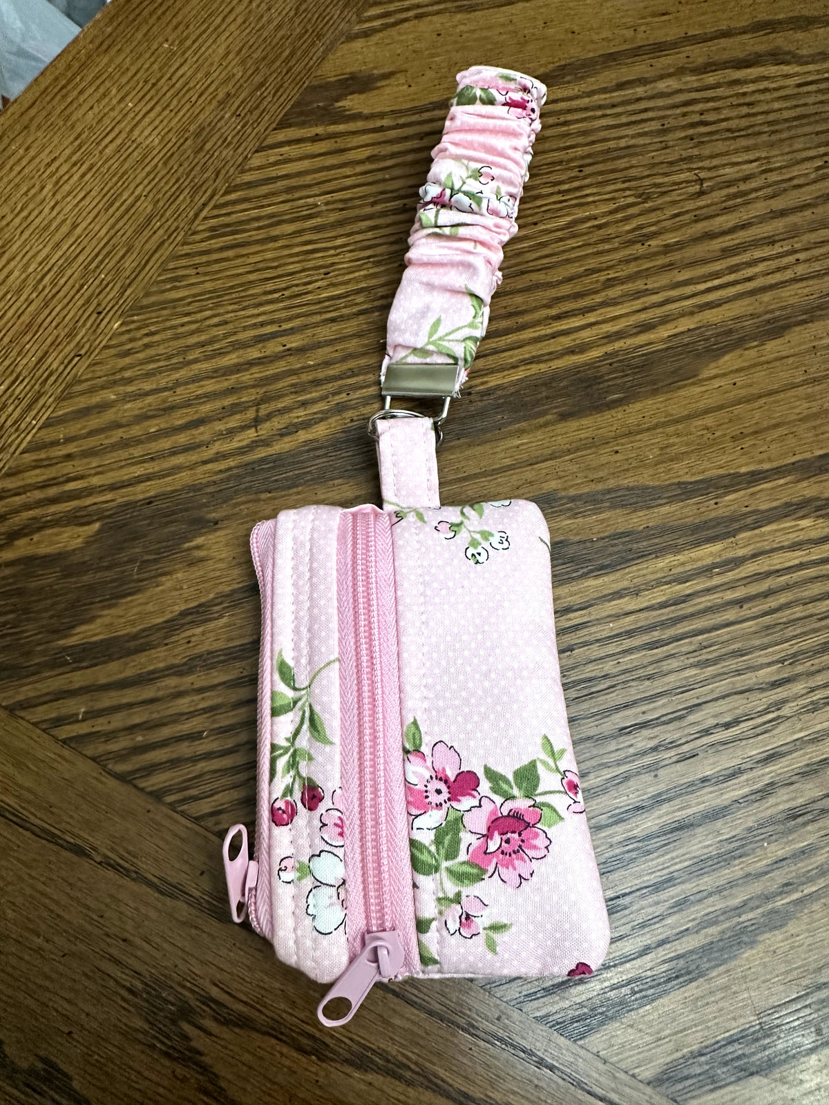 Front view of handcrafted pink floral fabric wristlet ID wallet showing the dual pink zippers, floral pattern, and ruched wrist strap on a wooden table.